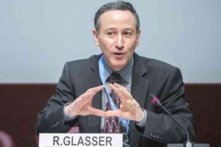 A man in a suit and tie speaks into a microphone at a conference table, seated behind a nameplate labeled "R. Glasser.
