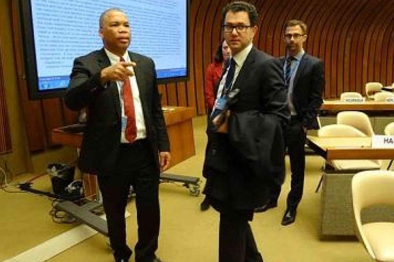 Four people in business attire stand and walk inside a conference room with empty chairs, nameplates, and a large presentation screen in the background.