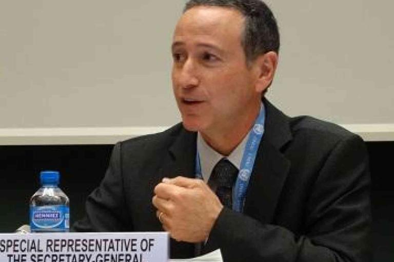 A man in a suit sits at a table with a microphone and a nameplate reading "Special Representative of the Secretary-General." A water bottle is also on the table.