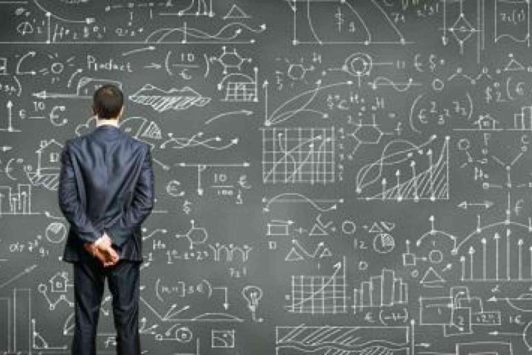 A man in a suit stands facing a large chalkboard filled with complex graphs, charts, and mathematical formulas.