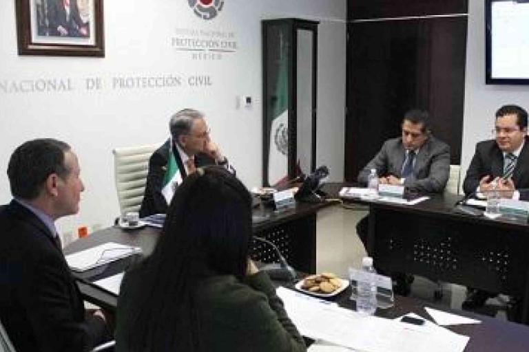 Five people in business attire sit around a conference table during a meeting in an office, with documents, water bottles, and snacks on the table.