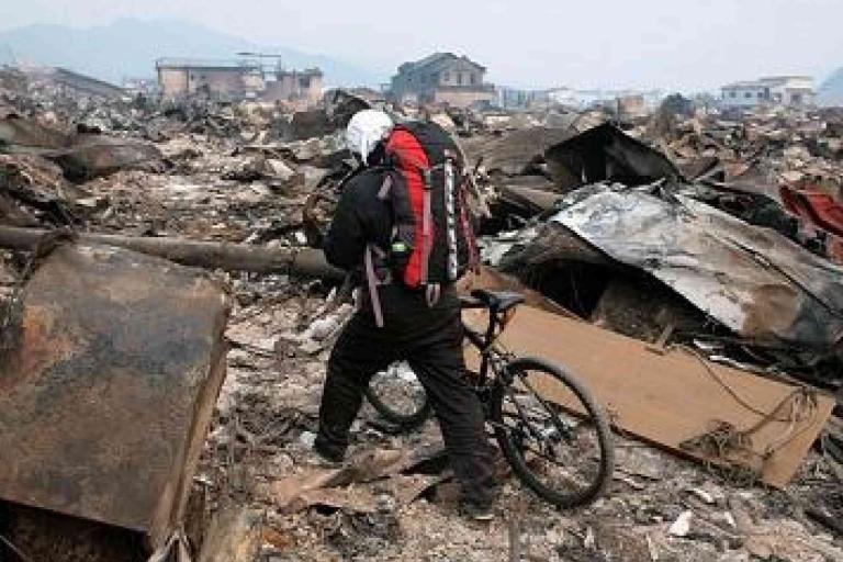 A person wearing a backpack and helmet walks a bicycle through debris and rubble in a devastated area with damaged buildings in the background.