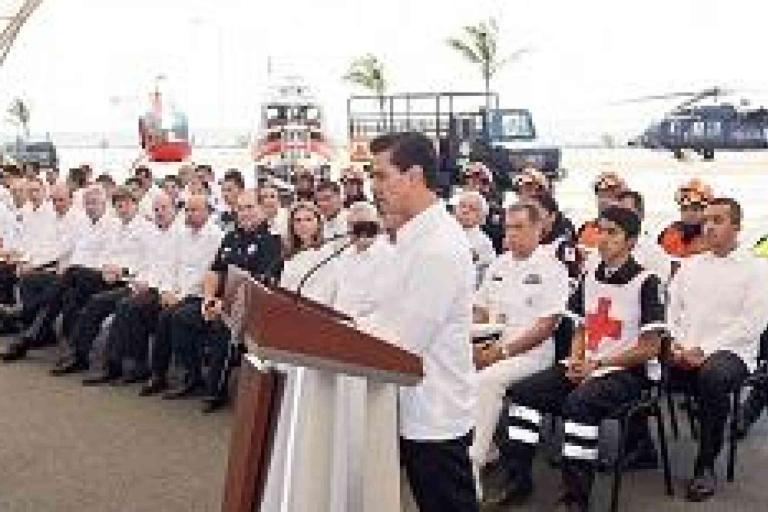 A man speaks at a podium in front of an audience of uniformed personnel, including police, Red Cross, and emergency responders, with vehicles and a helicopter in the background.