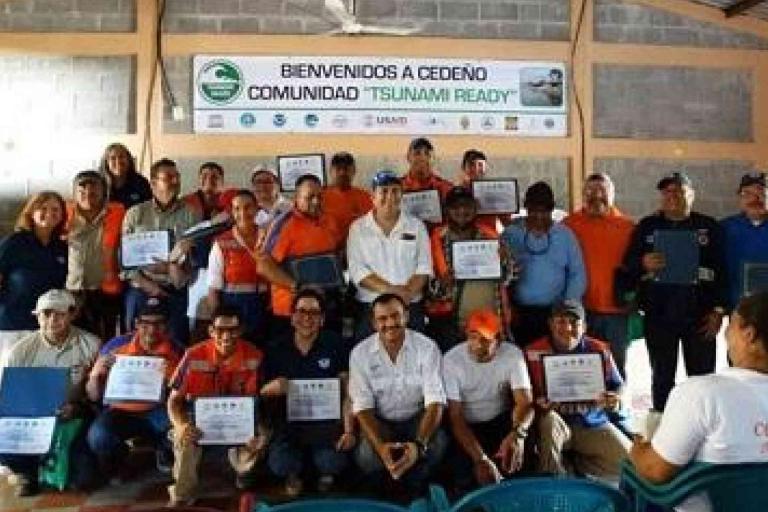 A group of people pose indoors holding certificates under a banner that reads “Bienvenidos a Cedeño Comunidad ‘Tsunami Ready’.”.
