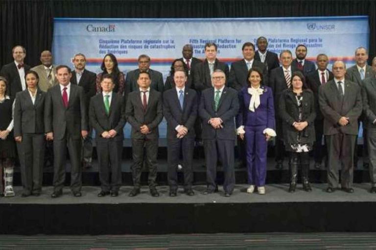 A group of formally dressed people pose for a photo on a stage with a blue banner in the background displaying Canadian and UN logos.