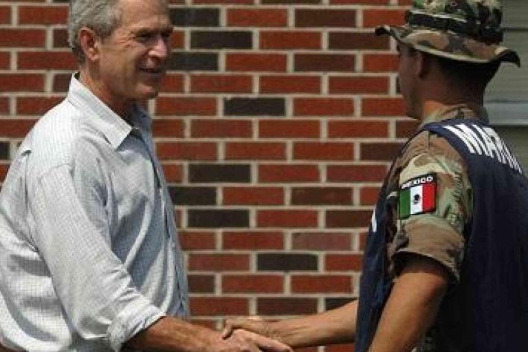 A man in a light shirt shakes hands with a uniformed soldier wearing a helmet and a vest labeled "MARINA" with a Mexican flag patch, in front of a brick wall.