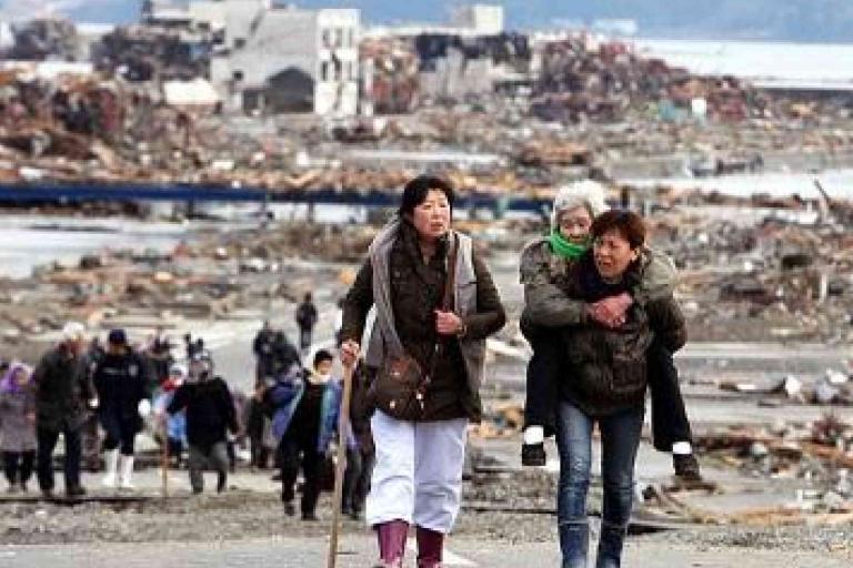A group of people walk through a devastated area filled with debris; one woman carries an elderly person on her back.