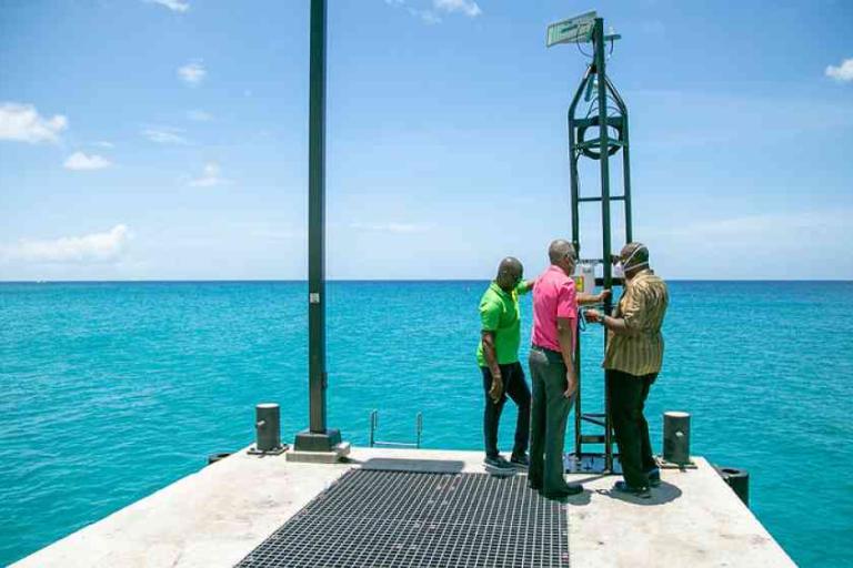 Three men stand on a pier next to a tall metal ocean monitoring device, with clear blue water and sky in the background.