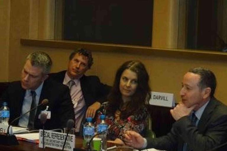 Four people sit at a conference table with nameplates and bottled water, engaged in discussion in a formal meeting room.