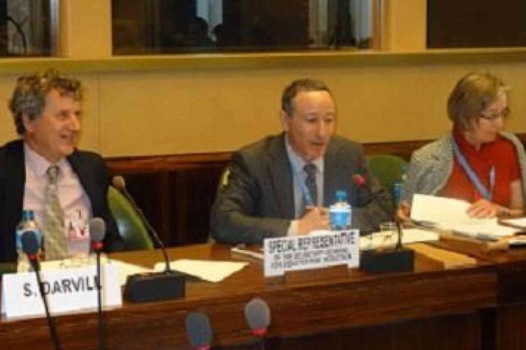 Three people sit at a conference table with microphones and documents; the man in the center speaks while the others listen. Nameplates are visible in front of each person.