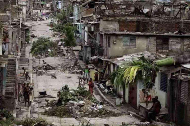 A street lined with damaged buildings, debris, and fallen branches, with people walking and sitting outside in a post-disaster urban area.