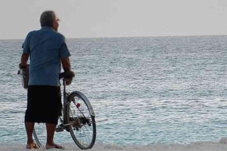 An older person stands barefoot on the beach holding a bicycle, looking out at the calm sea under a clear sky.