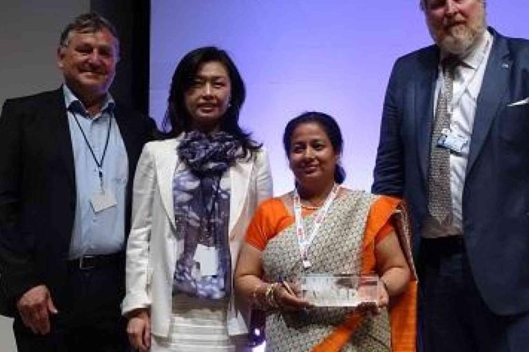Four people pose for a photo at an event; one person, dressed in an orange saree, holds a glass award.