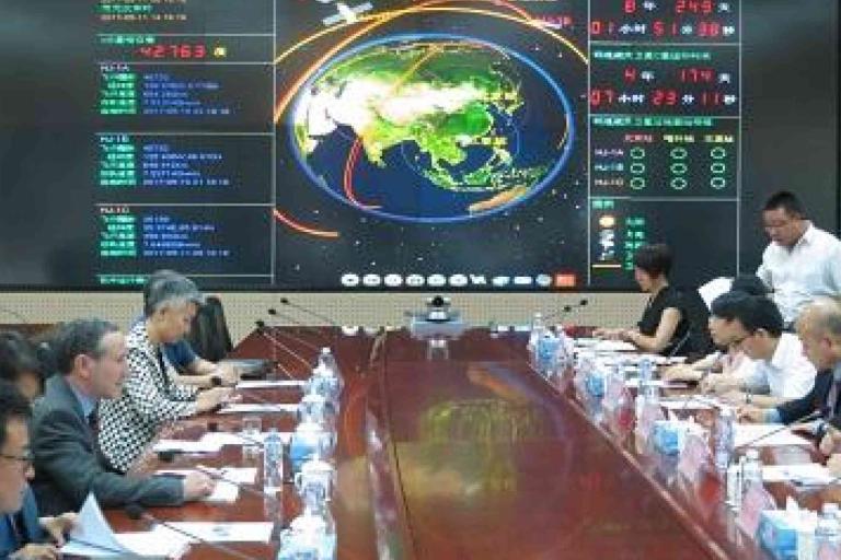 People seated on both sides of a conference table with documents, in front of a large digital screen displaying satellite data and a map of Asia.