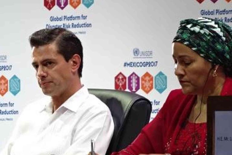 Two people sit at a table during the Global Platform for Disaster Risk Reduction event, with branded banners in the background. One person writes while the other looks forward.