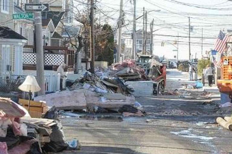 A residential street is lined with debris, damaged furniture, and trash piles, with utility poles and houses visible on both sides. People are in the background near an orange dumpster.