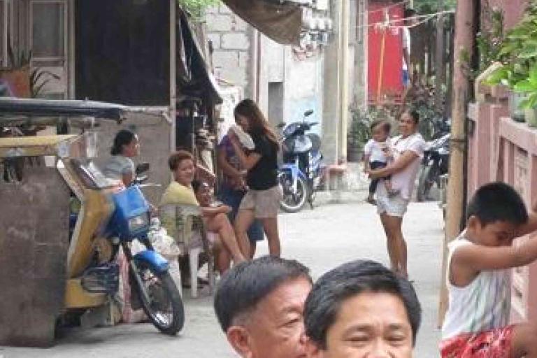 Several people, including children and adults, gather in a narrow urban alleyway with a tricycle, plants, and buildings visible in the background.