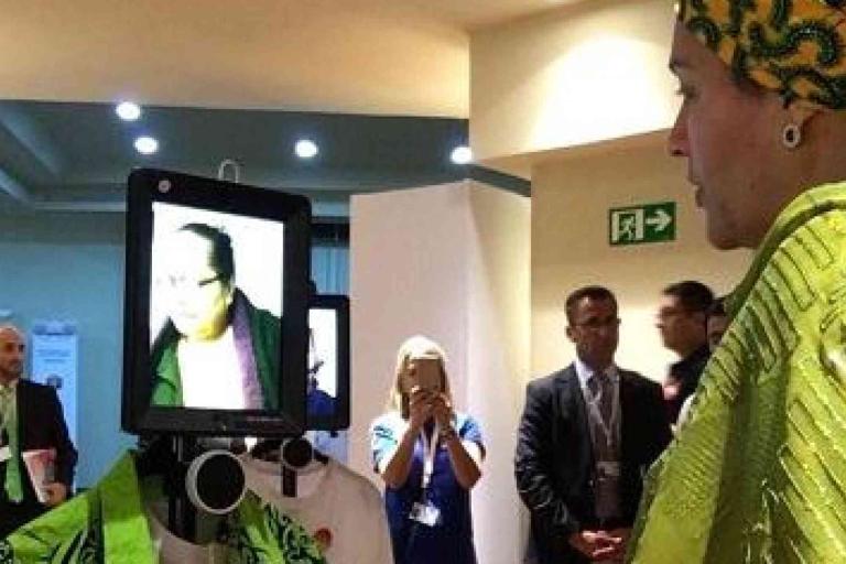 A person in traditional clothing interacts with a telepresence robot displaying another person's face on a screen, while several people observe in the background.