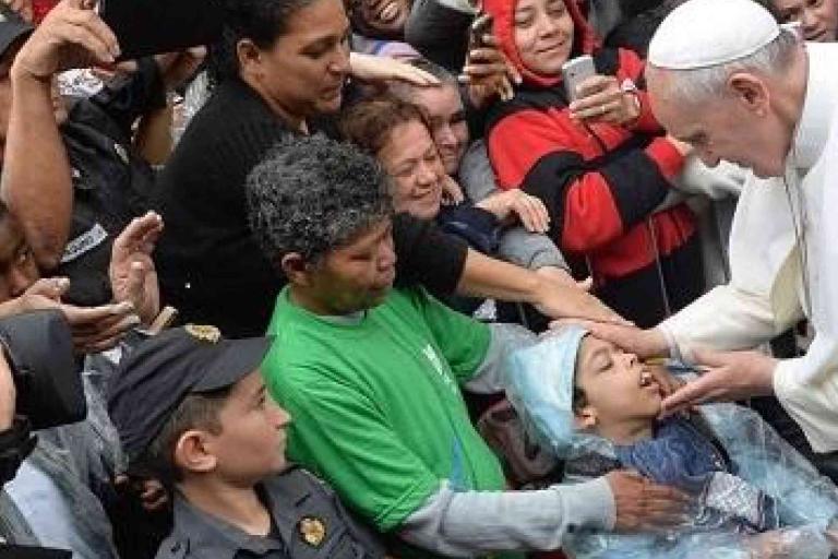 Pope Francis touches the face of a child in a wheelchair, surrounded by a crowd of people, some taking photos and police officers present.