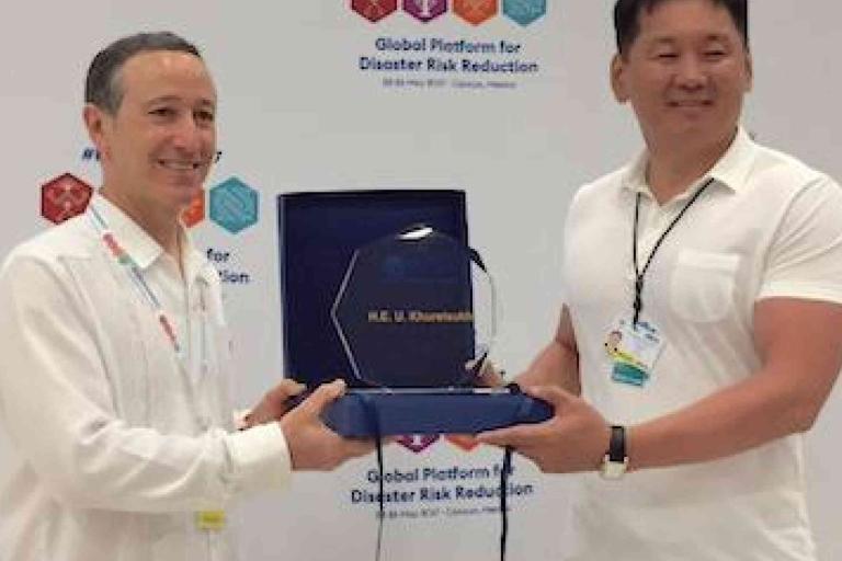 Two men stand together as one presents an award plaque to the other at an event for disaster risk reduction. Both are smiling and wearing white shirts.