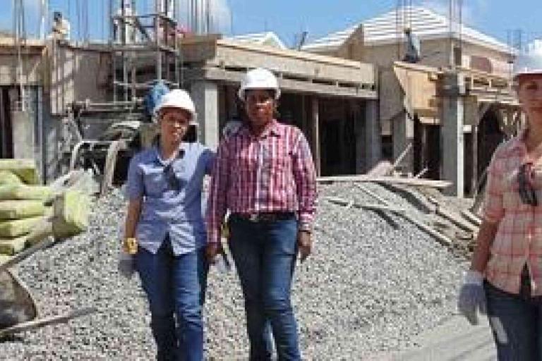 Three people wearing hard hats and gloves stand at a construction site with building materials and partially built structures in the background.