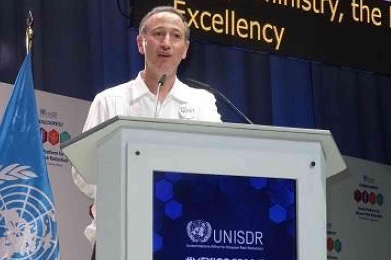A man stands at a podium with a UNISDR logo, speaking at a conference; a United Nations flag is visible beside him.