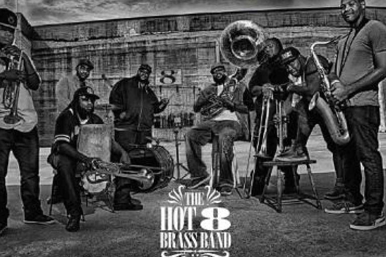 A group of eight musicians pose outdoors with brass instruments; the text at the bottom reads "The Hot 8 Brass Band.