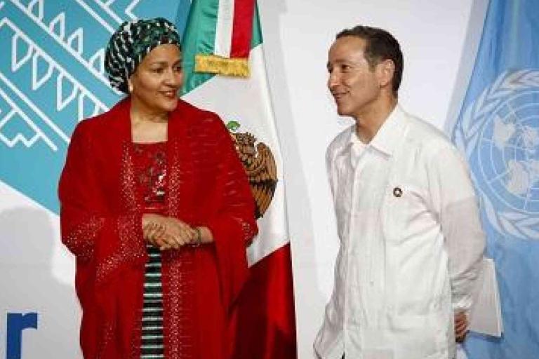 Two people stand in front of Mexican and United Nations flags, engaged in conversation at an official event.