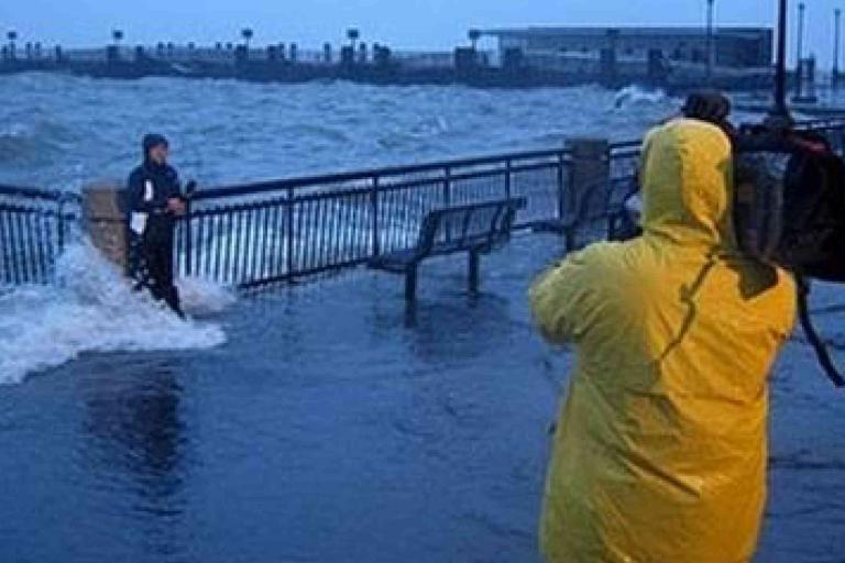 A person in yellow rain gear films a reporter standing by a flooded waterfront with waves crashing over a railing during stormy weather.