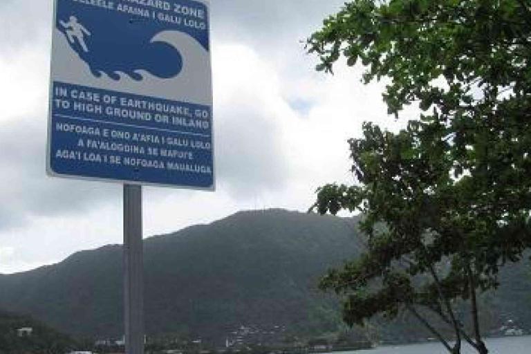 A tsunami evacuation zone sign instructs people to go to high ground or inland in case of an earthquake, with a coastal landscape and hills in the background.