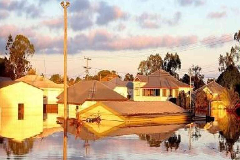 Residential neighborhood with houses partially submerged in floodwater, reflecting the buildings and sky, with trees and power lines in the background.