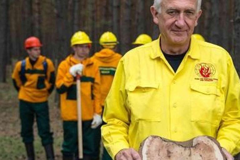 A man in a yellow uniform holds a slice of tree trunk, while four people in safety gear stand in the background in a forest.