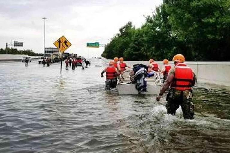 People wearing life vests wade through a flooded highway, some pulling a small boat with belongings, while others walk further ahead.