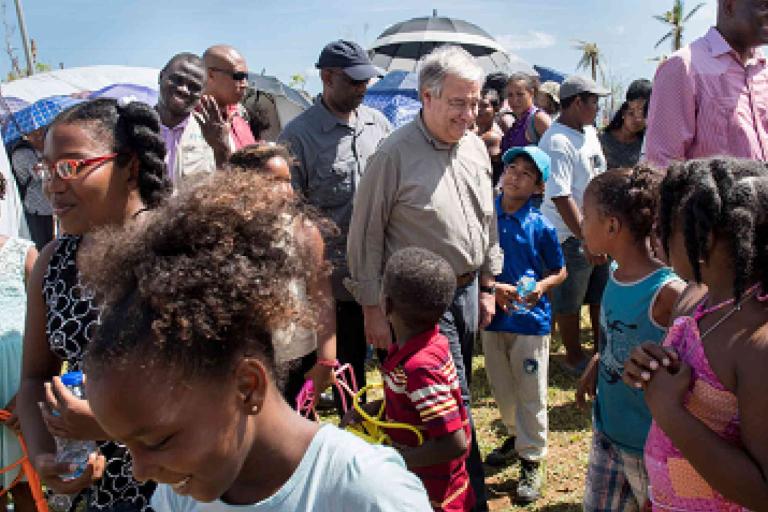 A group of adults and children gather outdoors near tents on a sunny day, some standing and some talking among themselves.