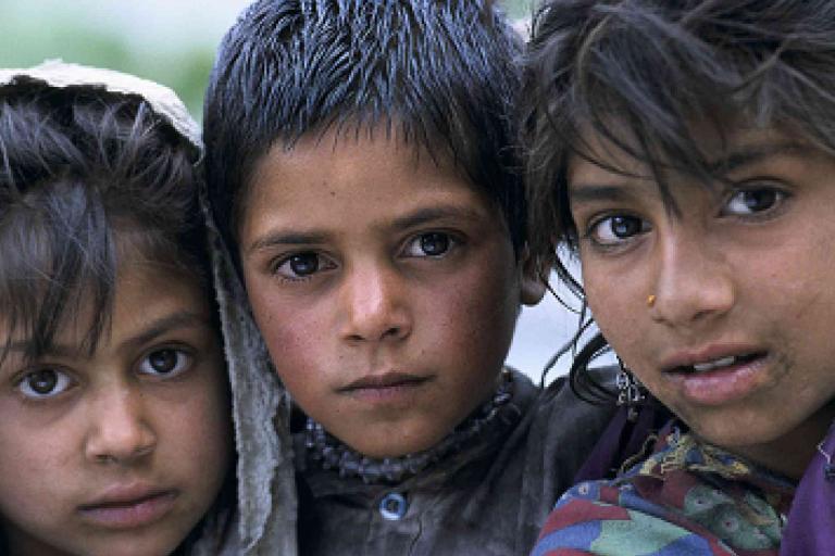 Three children with solemn expressions look directly at the camera. They have dark hair and wear simple clothing. The background is blurred.
