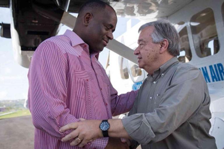 Two men greet each other and shake hands beside a small aircraft on a tarmac during daytime.