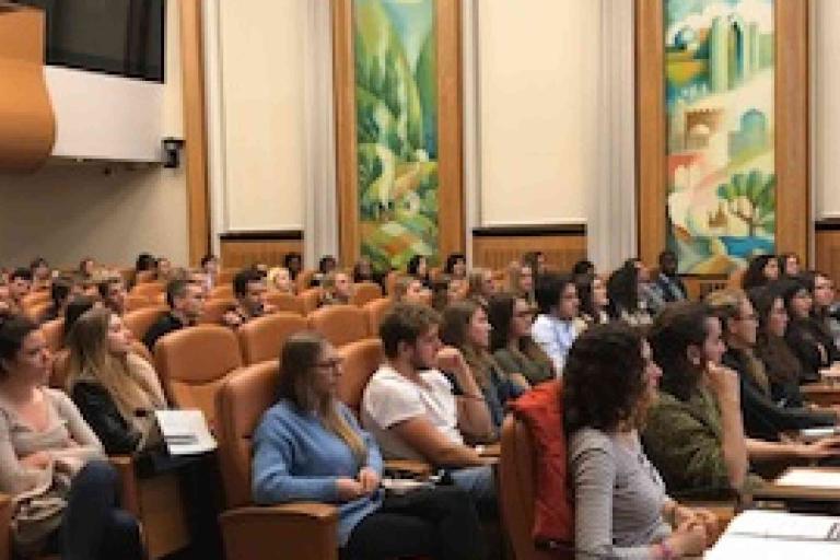 Audience seated in rows of tan chairs attentively listens to a presentation in a large room with decorative wall art and high ceilings.