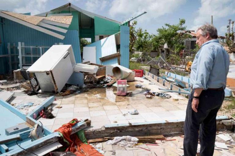 A man stands and observes severe damage to a house, with debris and household items scattered, after a natural disaster.
