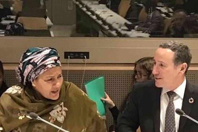 Two people sit at a conference table with microphones in front of them, appearing to be engaged in discussion, with others seated in the background.