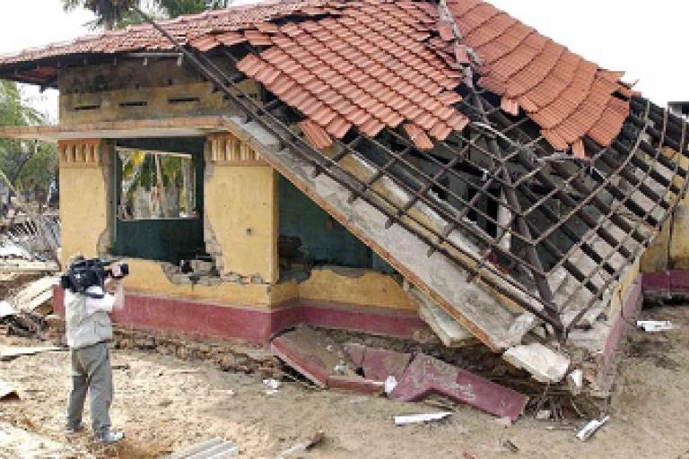 A damaged house with a collapsed roof; a person with a video camera films the scene amid scattered debris.