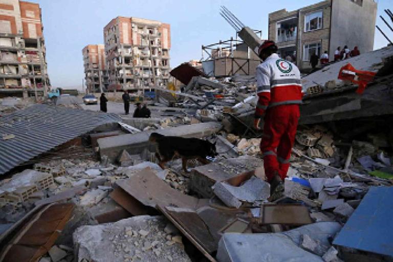A rescue worker walks over rubble and debris in a city area with collapsed and damaged buildings in the background.
