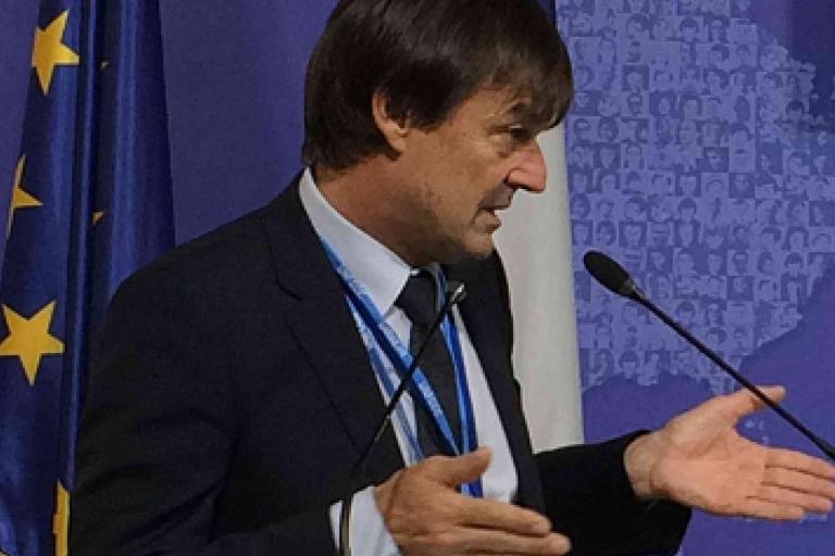 A man in a suit speaks at a podium with microphones, standing in front of an EU flag and a blue background.