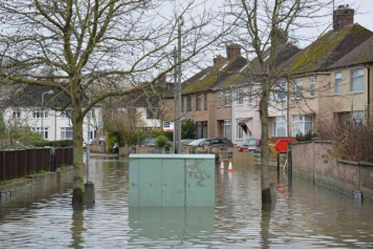 A residential street is flooded, with water covering the road and reaching up to house entrances. Trees and parked cars are partially submerged.