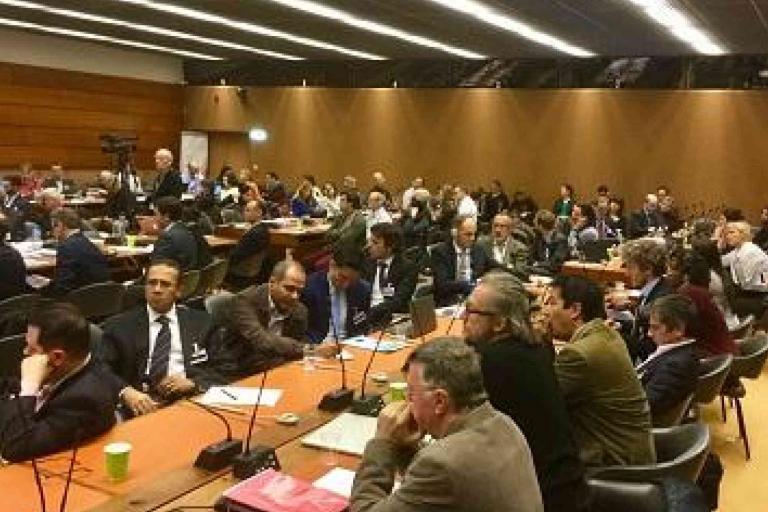 A large group of people sit at tables in a conference room, engaged in discussion during a formal meeting or event.