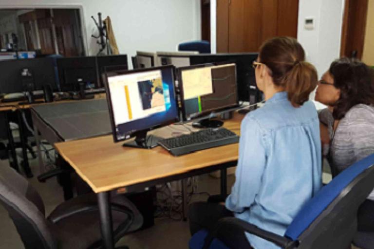 Two people sit at a desk in an office, looking at maps and data displayed on dual computer monitors.