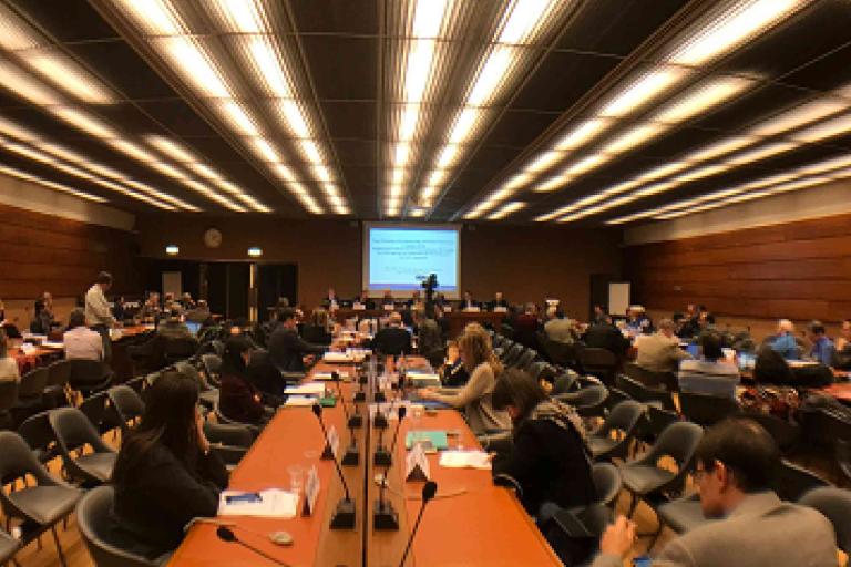 A large conference room filled with people seated at long tables, facing a presentation screen at the front of the room.