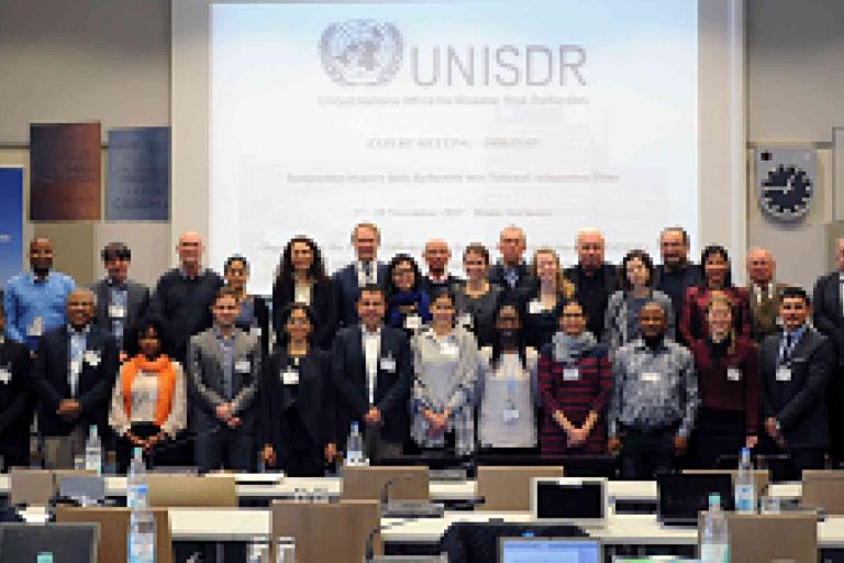 A group of people pose for a photo in a conference room with a UNISDR presentation projected on the screen behind them.