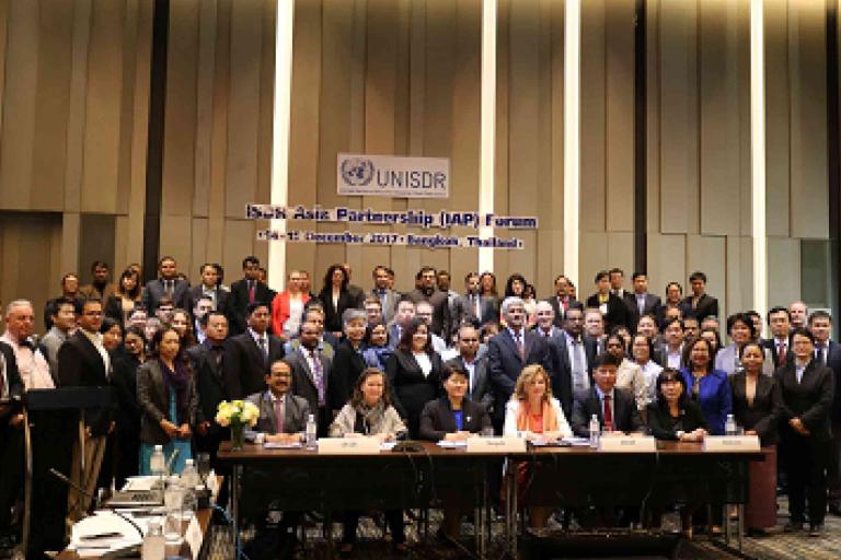 A large group of people in formal attire pose for a group photo at the UNISDR Asia Partnership Forum in a conference room with a sign reading "Bangkok, Thailand" in the background.