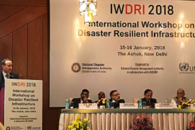 A speaker addresses attendees at the IWDRI 2018 International Workshop on Disaster Resilient Infrastructure in New Delhi, with panelists seated at a table.