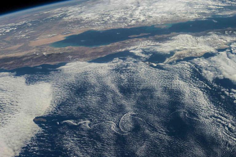 View of Earth's surface from space showing land, ocean, and cloud formations with a portion of the planet's curvature and atmosphere visible.